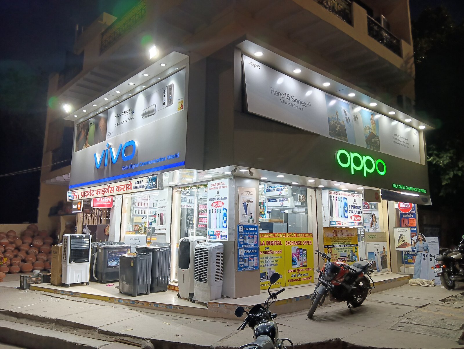 A corner mobile phone shop in Dabri, Delhi, lit up at night with phone brand signboards and finance offer posters