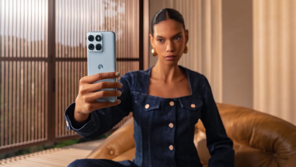 A woman holds up the Motorola Edge 70 Fusion in a pale blue finish, camera side facing out, in a bright living room