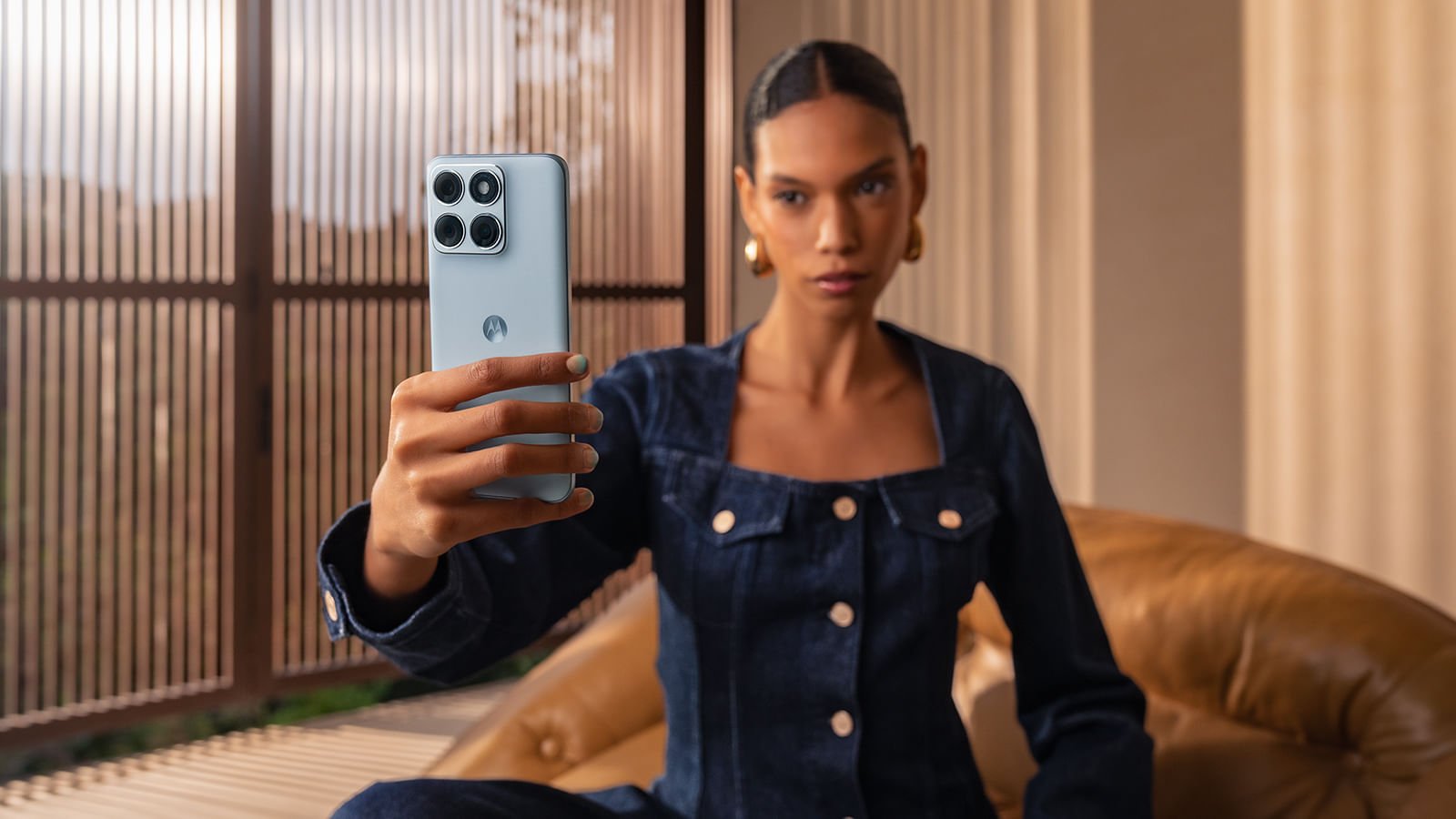 A woman holds up the Motorola Edge 70 Fusion in a pale blue finish, camera side facing out, in a bright living room