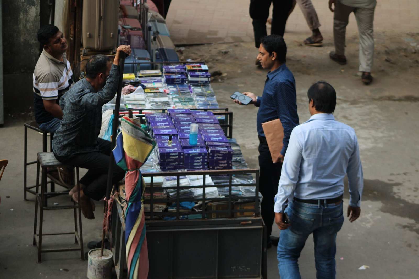 A man checks a phone beside a street stall piled with phone cases and screen guards, with a stallholder and passers-by